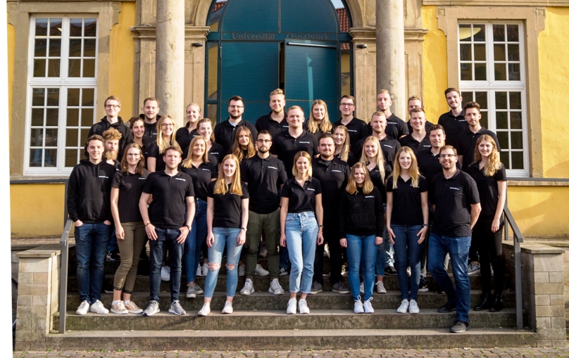 Gruppenfoto der Mitglieder der Fachschaft Wirtschaftswissenschaften (fachschaft|wiwi) aus dem Jahr 2018 auf den Treppen des Haupteingangs vom Schloss Osnabrück. Alle 34 Personen tragen ein schwarzes Polo-Shirt mit dem Logo der Fachschaft bzw. einen schwarzen Hoodie.